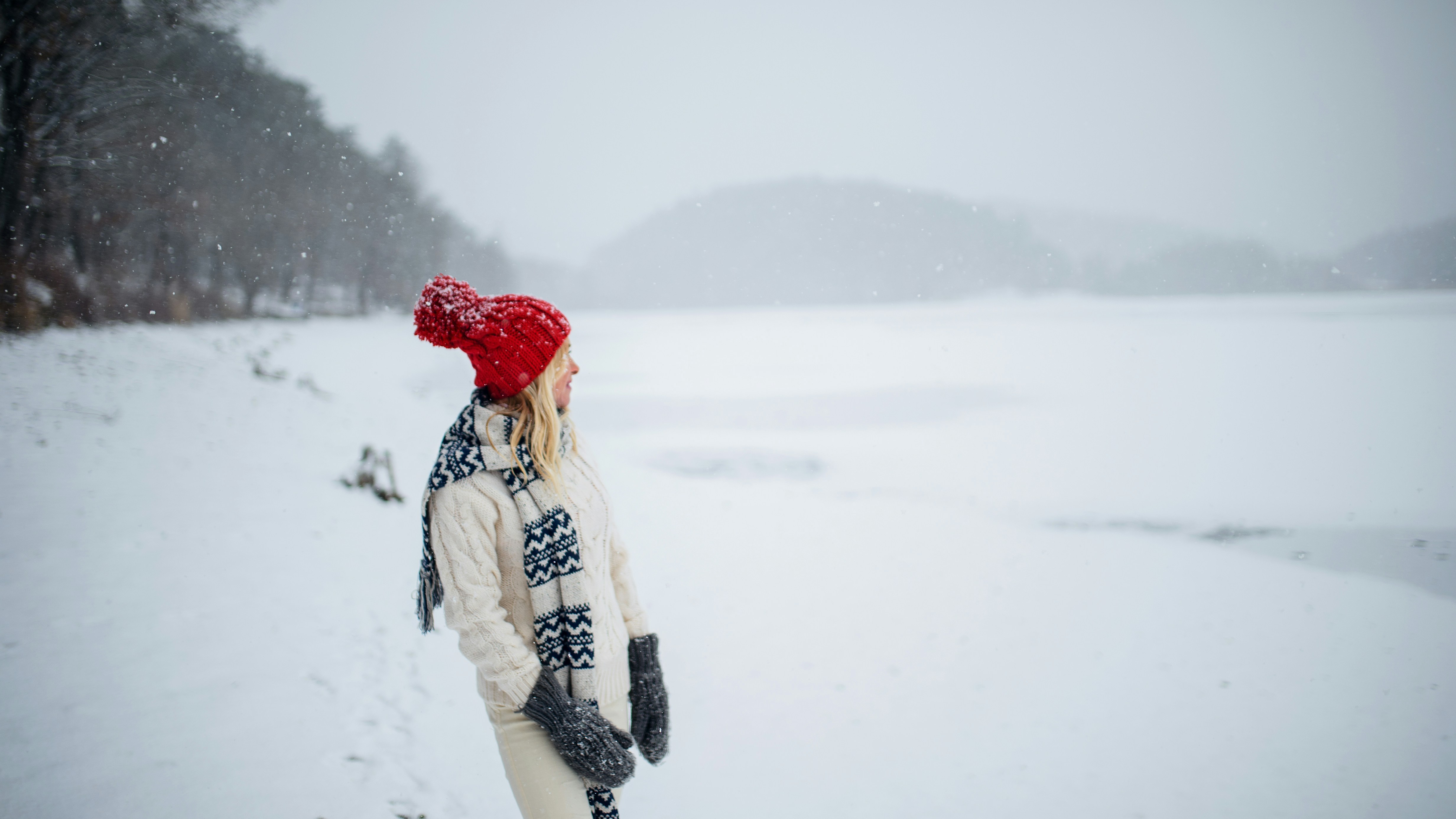A person in a red hat, a white sweater, and a black and white chevron scarf stands in a snowy landscape by a frozen lake, looking out at the hills.