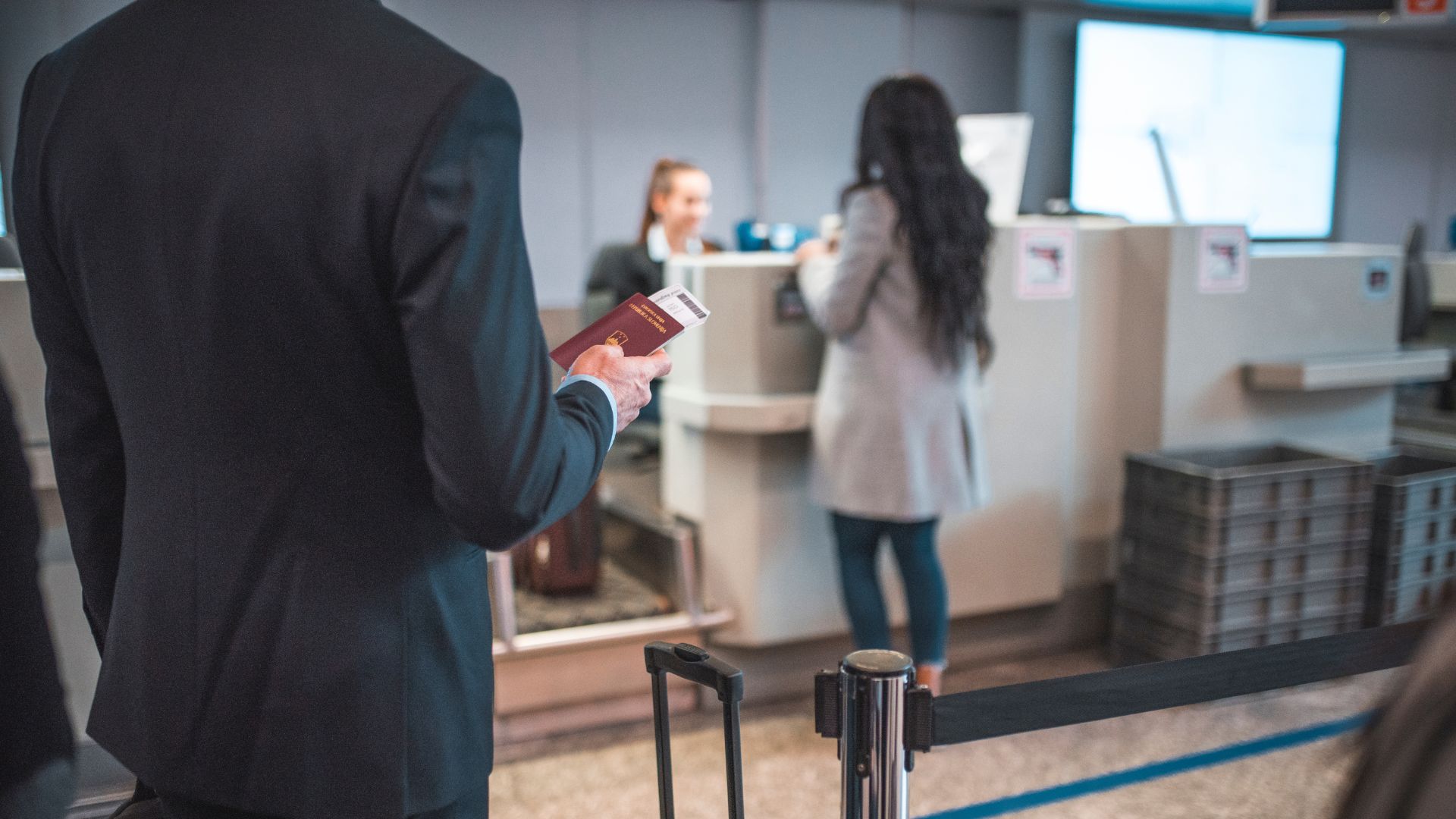 A stock photograph of passengers at a generic airport check-in desk, where a man in a black suit holds a passport and boarding pass, and a woman in a gray coat is interacting with the check-in agent.