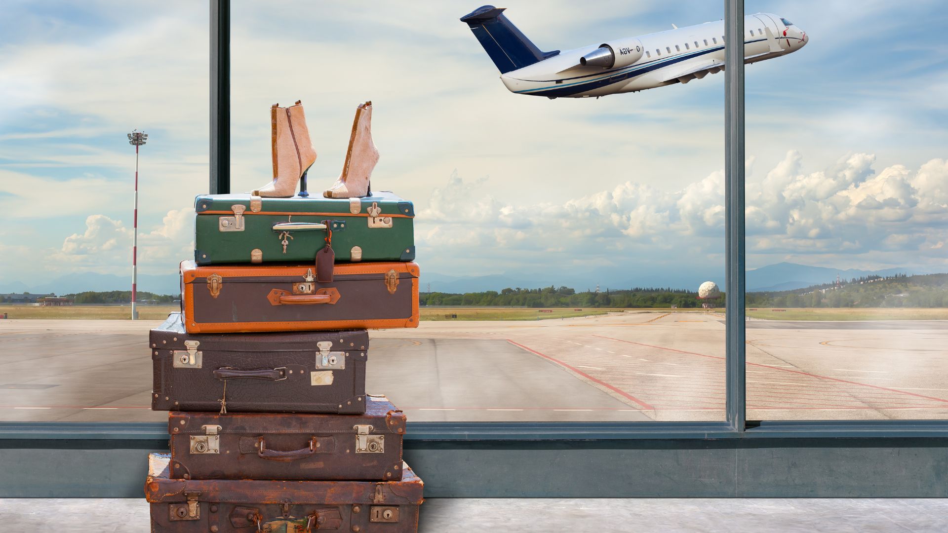 A stock photograph of a stack of vintage-style suitcases in an airport terminal with large windows looking out onto the tarmac at a private jet in the sky at sunset.