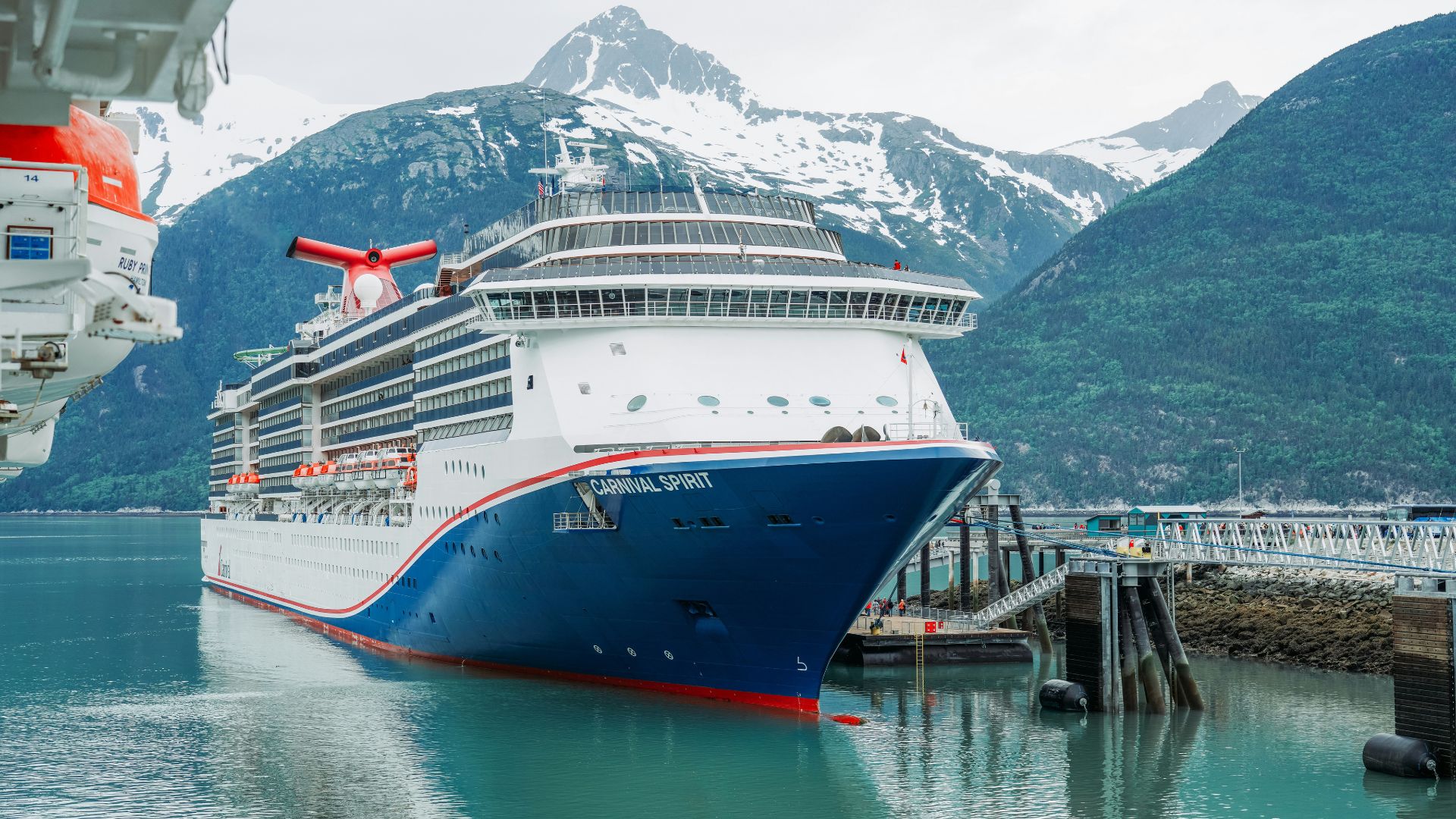 The large cruise ship Carnival Spirit docked in an Alaskan port surrounded by steep, green and snow-capped mountains.