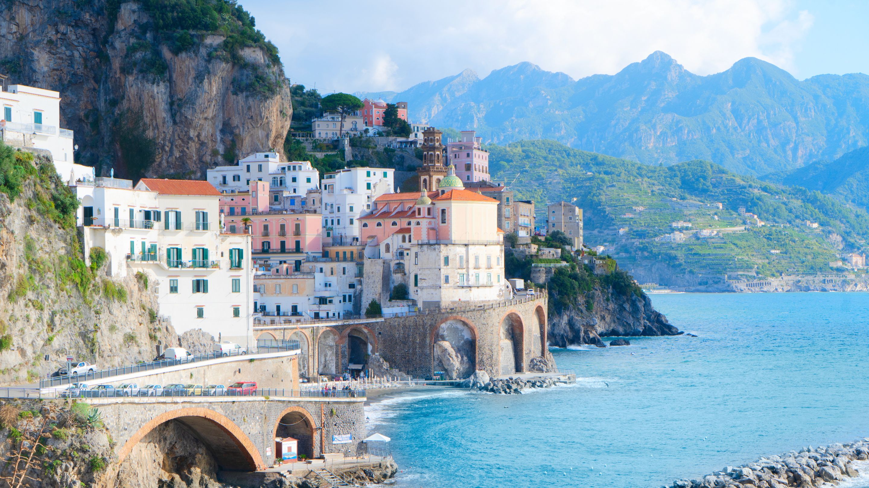A wide view of the picturesque Italian fishing village of Atrani, showing its pastel-colored buildings and a large stone viaduct built over the beach and leading into a mountain cliff, all set against a vibrant blue Mediterranean sea and green mountains.
