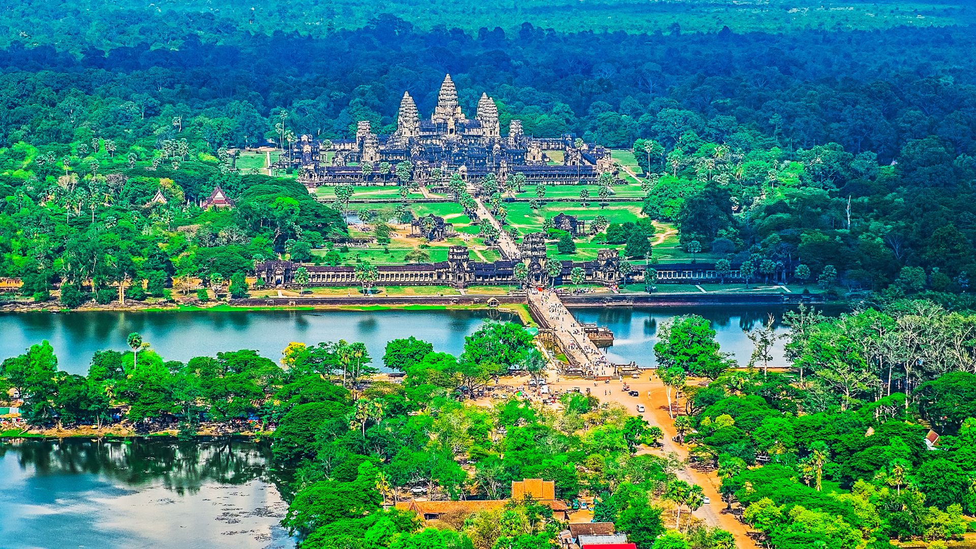 An aerial view of the ancient, sprawling Angkor Wat temple complex with its iconic five towers, surrounded by a large moat and lush green forest in Cambodia.