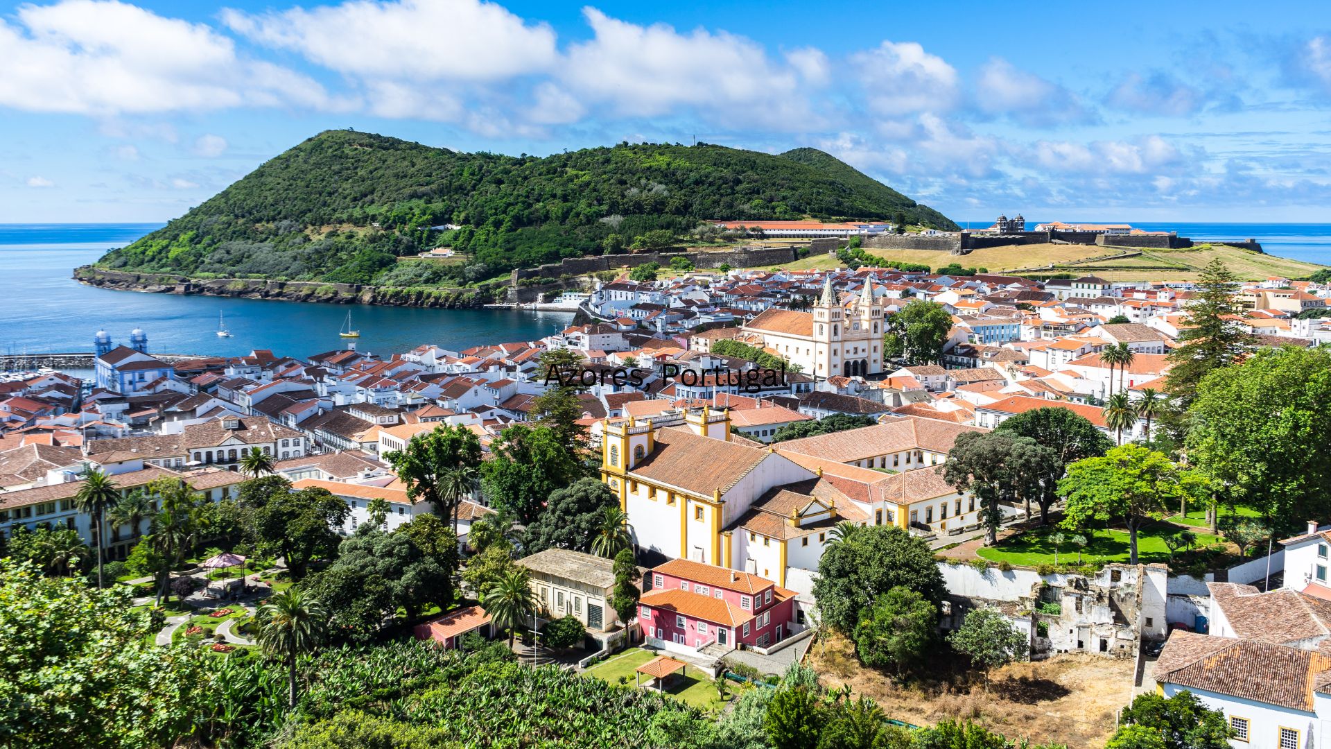 A panoramic daylight view of the historic, red-roofed city of Angra do Heroísmo on Terceira Island, Azores, with a large green volcanic hill (Monte Brasil) and the Atlantic Ocean in the background.