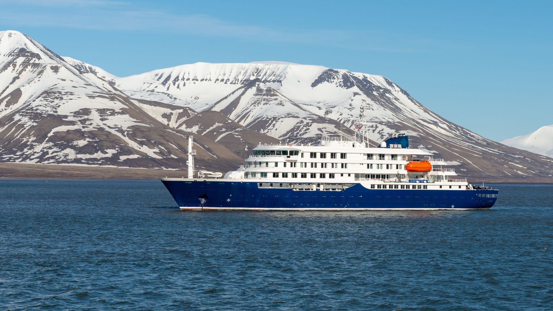 A large, blue and white expedition cruise ship, the MV Hondius, sails on dark blue water with large, snow-capped mountains and a blue sky in the background.