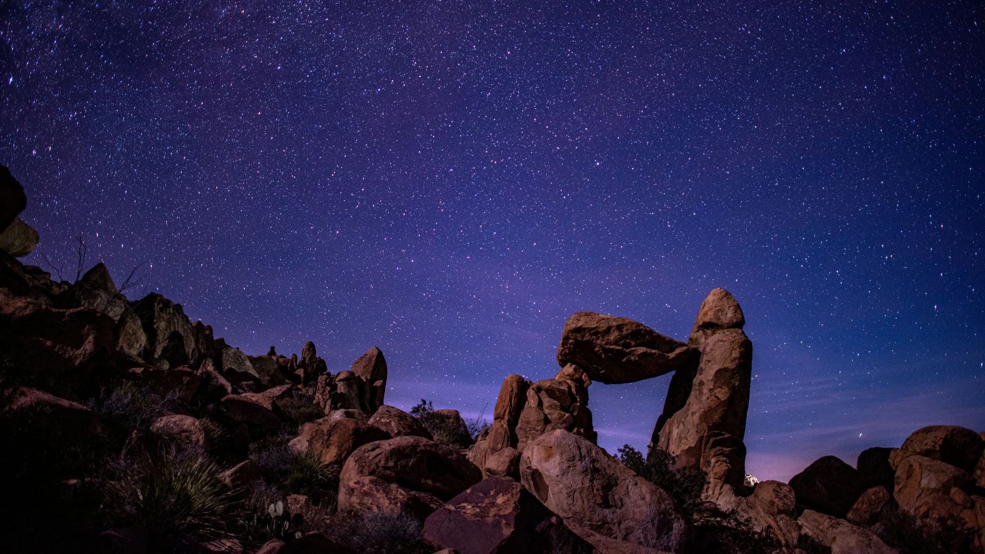 A nighttime long-exposure photograph of the "Balanced Rock" formation in Big Bend National Park, Texas. The unique rock arch is silhouetted against a deep blue and purple night sky densely filled with thousands of stars and the faint glow of the Milky Way galaxy. The foreground is dark and rocky, with desert vegetation visible around the large boulders.