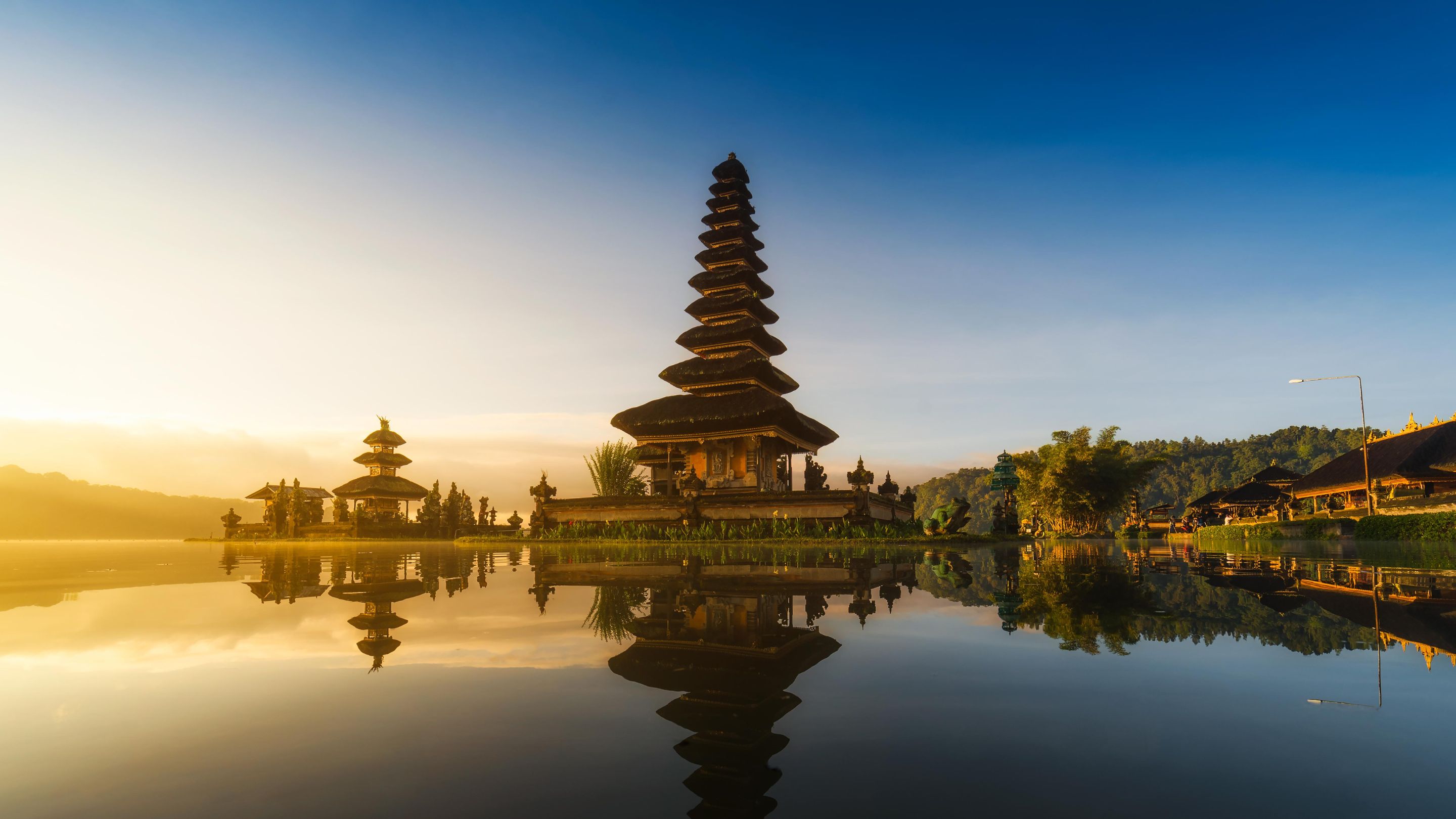 A wide-angle, sunrise view of the multi-tiered Balinese Hindu Ulun Danu Beratan temple on the edge of Lake Bratan, with the temple and surrounding buildings perfectly mirrored in the still water and mountains in the misty background.