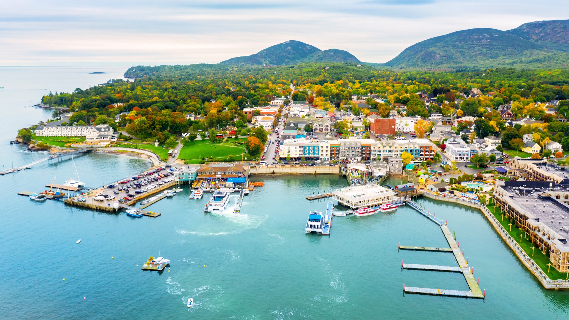 An aerial daytime view of the town of Bar Harbor, Maine, featuring a busy harbor with boats and docks in the foreground, with numerous colorful buildings and lush, tree-covered mountains in the background.