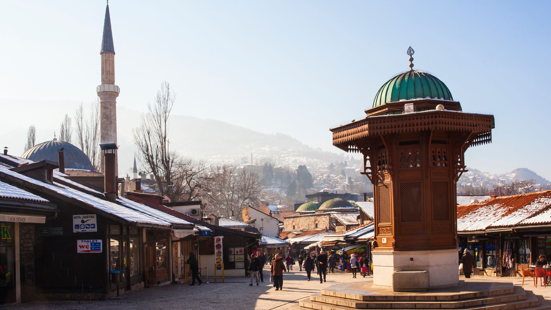 A bright, sunny photograph of the historic Baščaršija square in Sarajevo, Bosnia and Herzegovina, featuring the iconic octagonal wooden Sebilj fountain in the foreground and the minaret of the Baščaršija Mosque in the background, surrounded by snow-dusted market stalls.