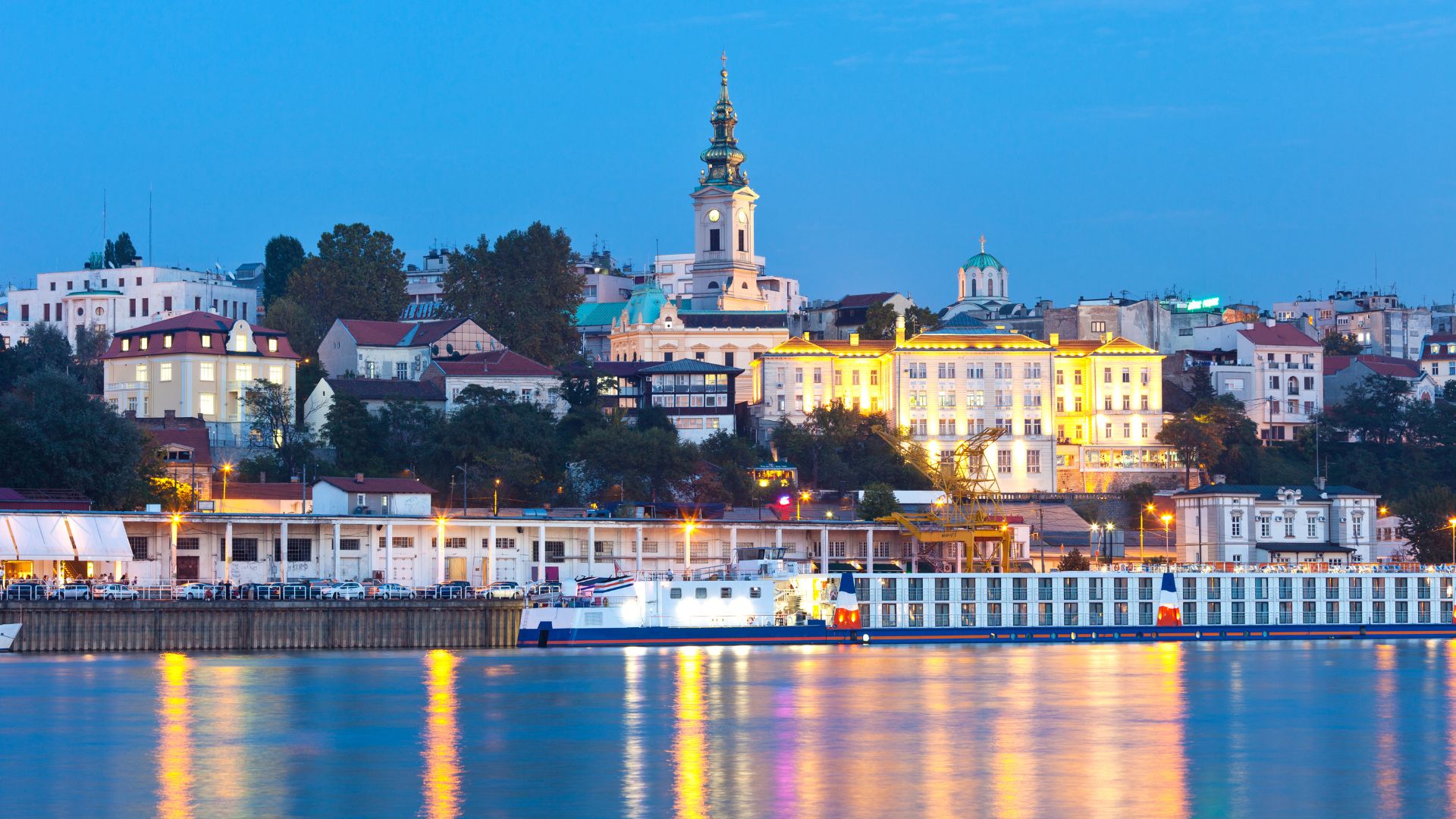 A twilight photograph of the Belgrade, Serbia, waterfront skyline, featuring brightly lit historic buildings and a prominent church bell tower, with colorful reflections shimmering on the river in the foreground.