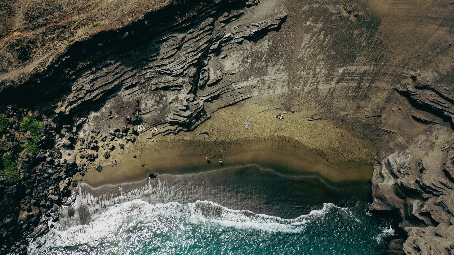 An aerial photograph of a remote green sand beach (Papakōlea Beach) on the Big Island of Hawaiʻi, nestled within a bay with high, rugged volcanic cliffs, and dark ocean waves breaking on the shore.