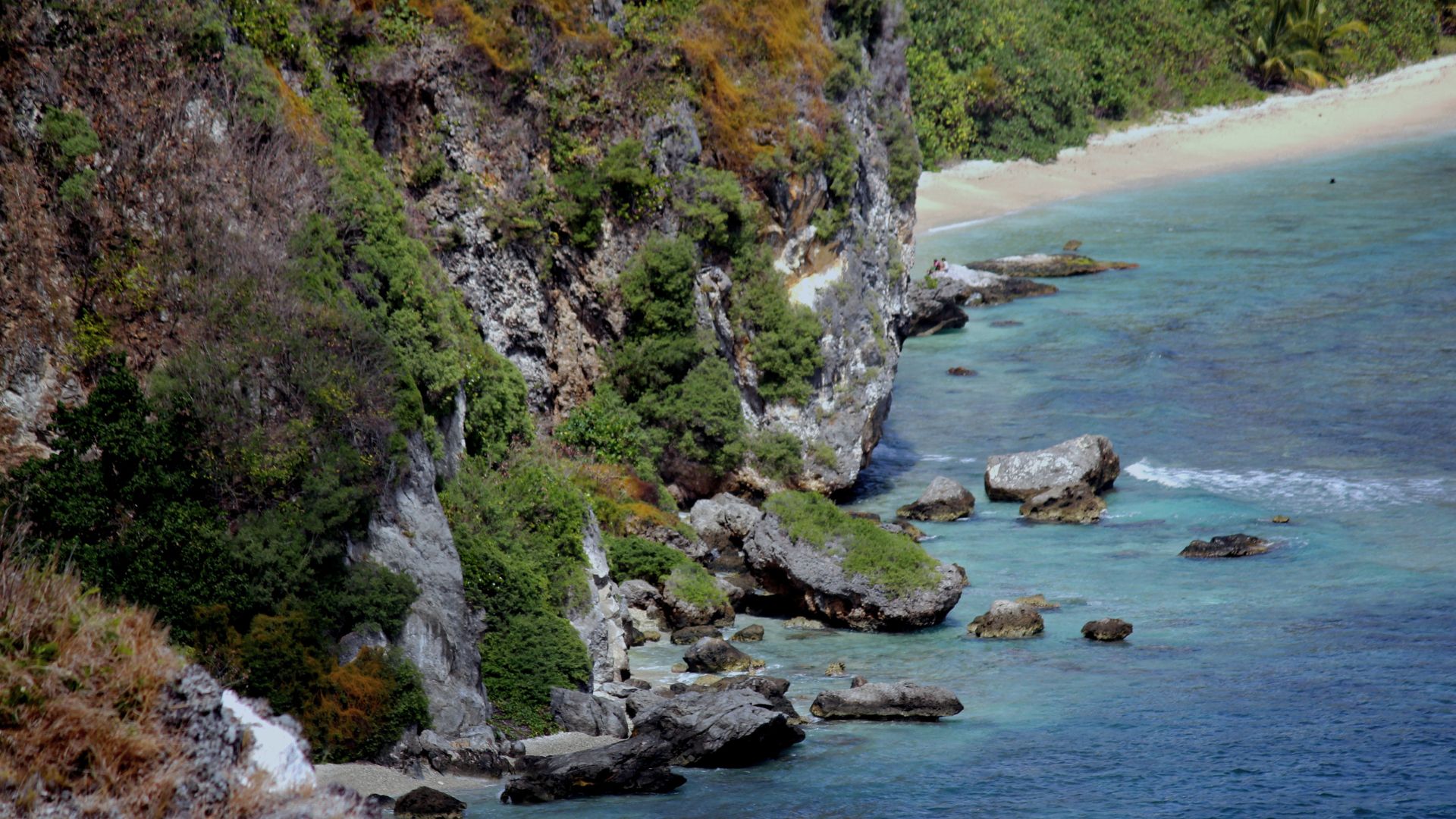 An elevated photo of a tropical coastline with a sheer, rocky cliff covered in lush green vegetation on the left, a small sandy beach in the distance, and turquoise ocean water dotted with large rocks.