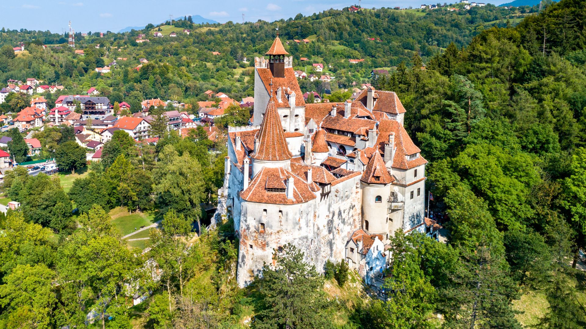 A medieval Gothic-style castle perched dramatically on a rocky cliff, with multiple red-tiled towers and spires, surrounded by a dense green forest and a small village in the valley below.