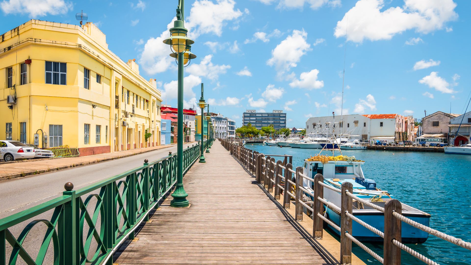 Daylight photo of a wooden boardwalk with a green railing next to a canal filled with boats, leading toward a yellow colonial building in Bridgetown, Barbados.
