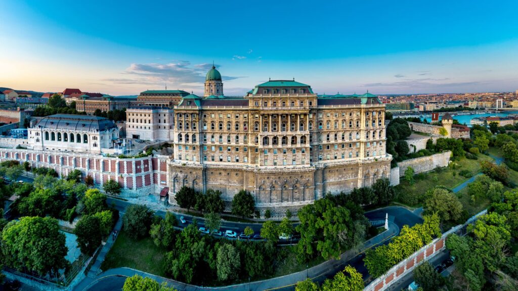 A sprawling, elevated view of the historic Buda Castle (Royal Palace) in Budapest, Hungary, situated on a lush green hill overlooking the city.