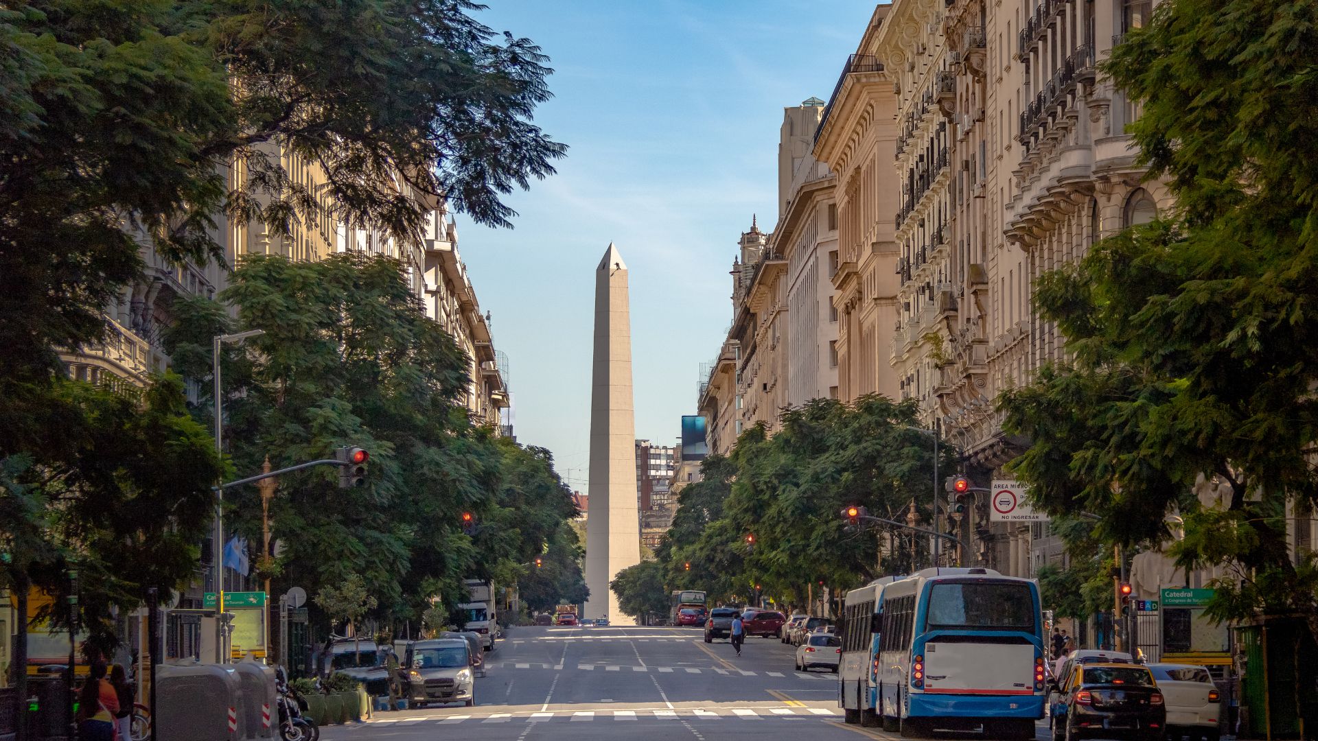 A wide, sunlit street view looking down a busy avenue lined with trees and tall, classic European-style buildings, with a tall, white stone obelisk monument standing prominently in the distance at a major intersection.