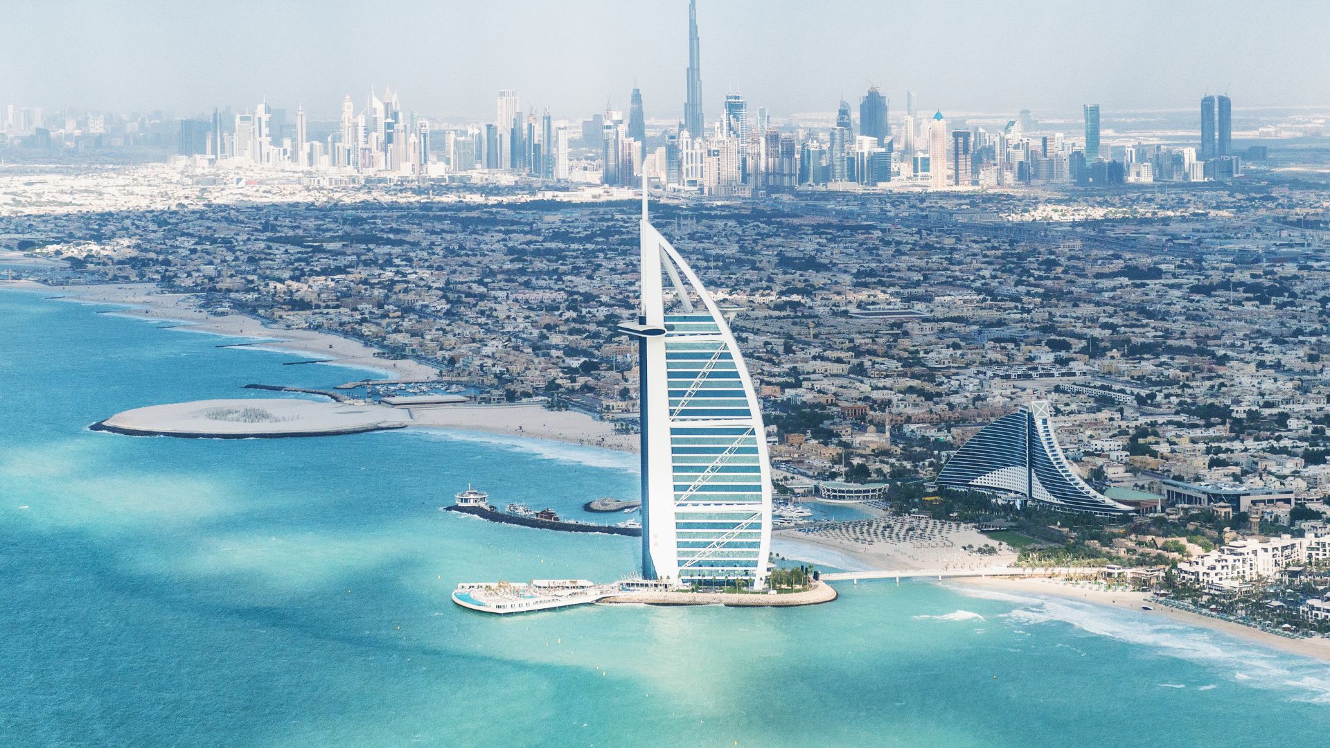 An aerial panoramic view of the Dubai coastline featuring the sail-shaped Burj Al Arab hotel on an artificial island, with other iconic skyscrapers and the city skyline in the background.