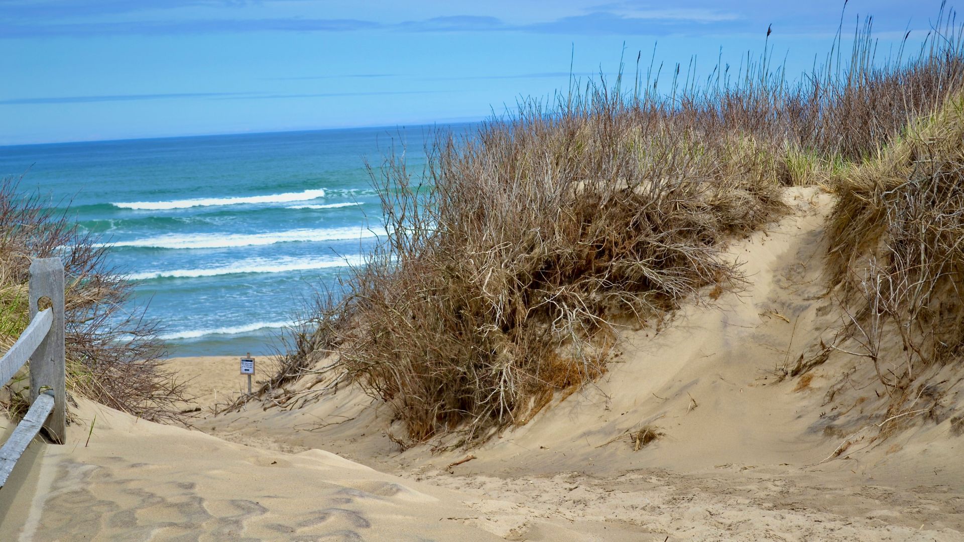 A sandy path and a low wooden fence lead down a large sand dune covered in dry beach grass toward the blue ocean with white breaking waves under a light blue, cloudy sky.