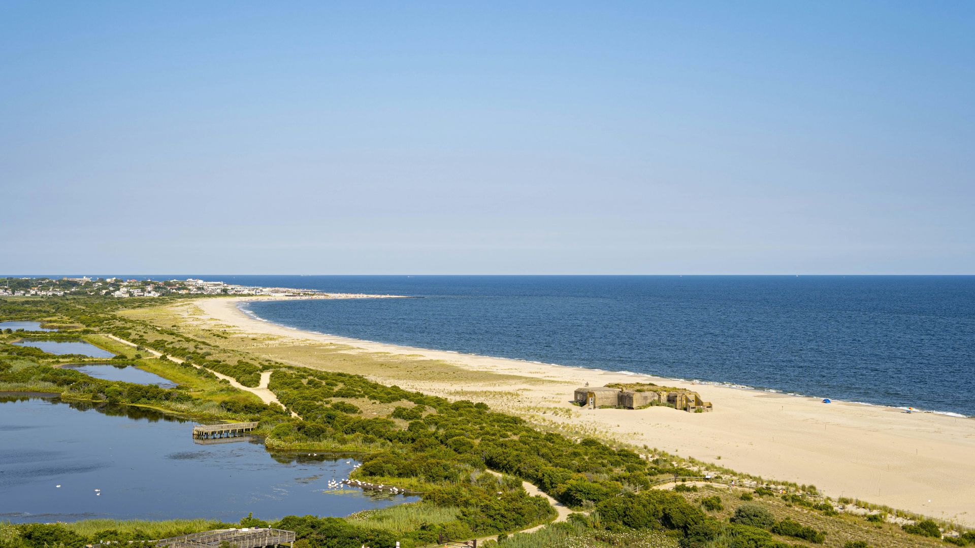 An elevated view of a sandy beach and coastal marshland with a concrete military bunker in the foreground, ocean in the distance, and a town visible on the far left of the coastline under a clear blue sky.