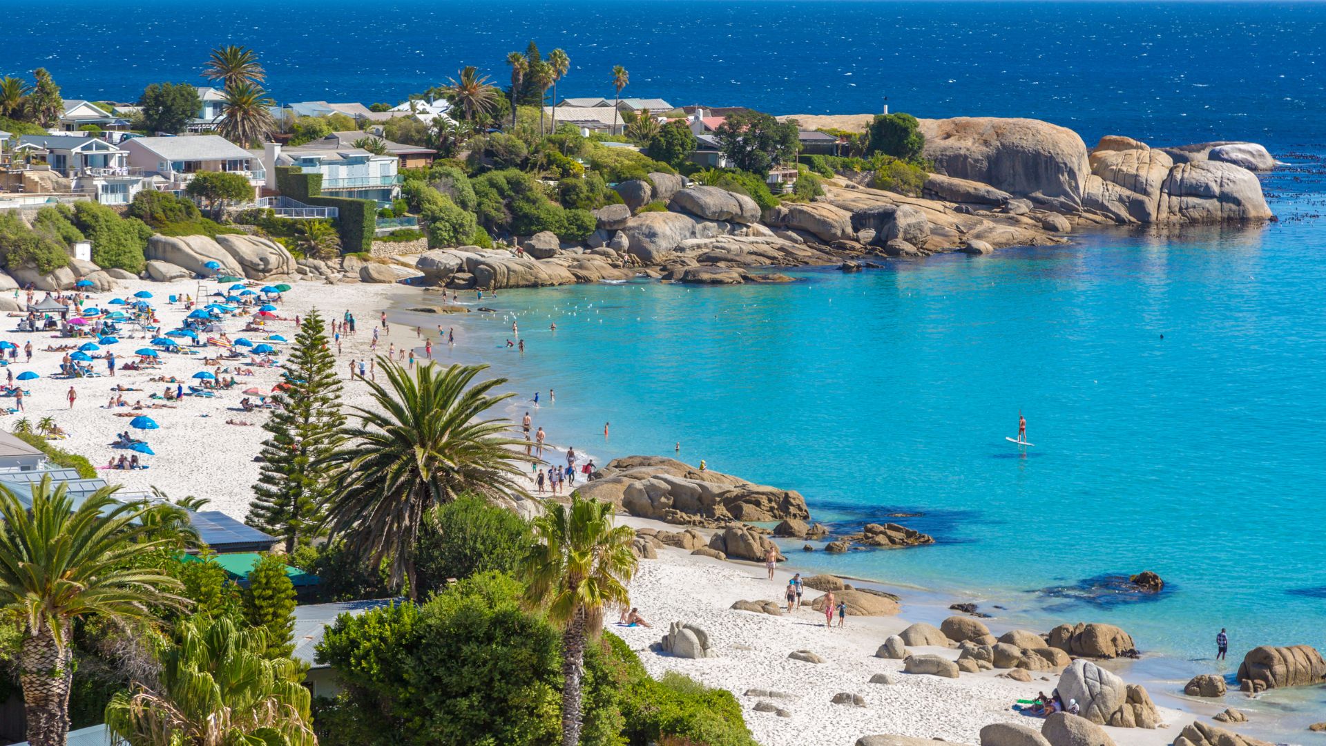 An aerial, sunlit view of a white sandy beach with turquoise water, palm trees, colorful beach umbrellas, people relaxing, and large granite rock formations and houses lining the coast.