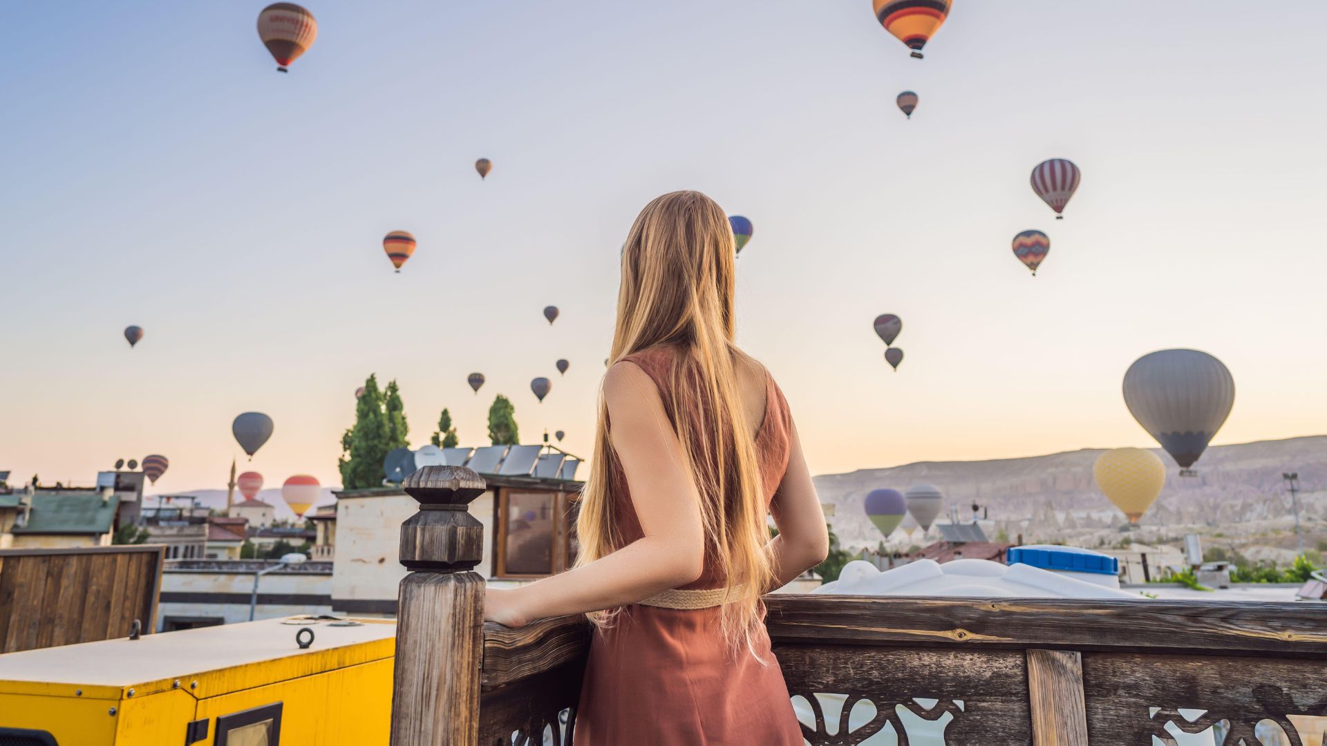 A woman with long blonde hair, viewed from behind, stands on a wooden terrace railing and looks out at a stunning sunrise sky filled with dozens of colorful hot air balloons floating over the unique rock formations and rooftops of Cappadocia, Turkey.