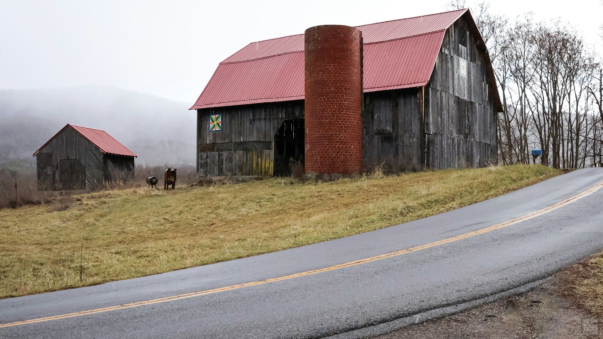 A landscape photograph of a large, weathered wooden barn with a red tin roof and an attached brick chimney sitting on a grassy hillside next to an asphalt road. A smaller wooden shed stands to the left, and two dark animals, likely cows or llamas, graze in the field. Hills covered in a light fog are in the background under a cloudy sky.