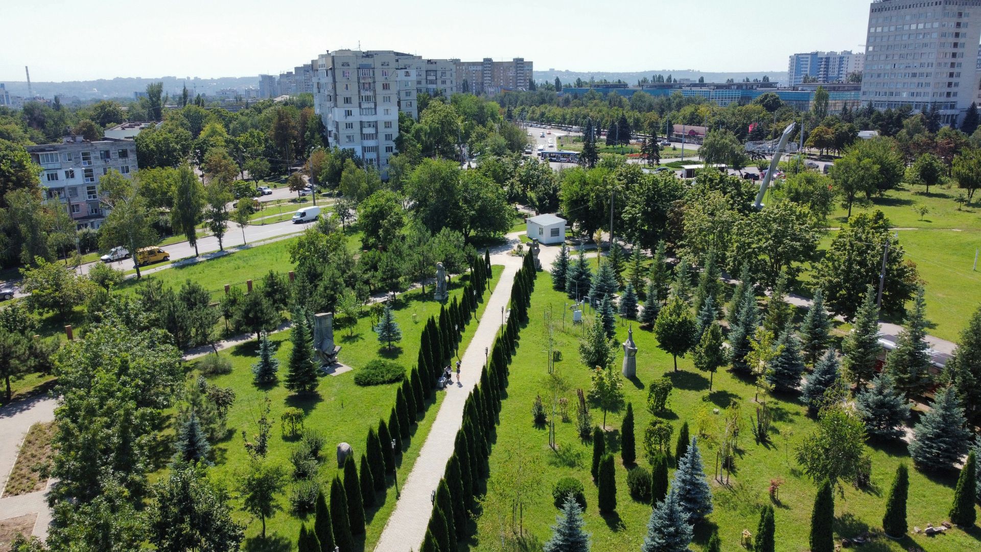 An aerial, daytime photograph of a lush green city park featuring a long, paved path lined with cypress trees and statues, leading toward a small white structure, with large residential apartment buildings visible in the background.