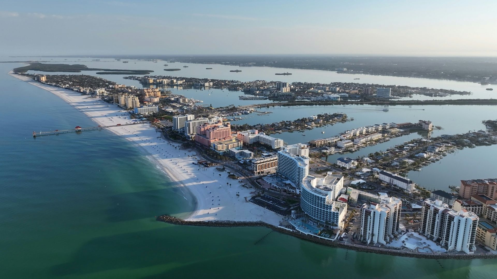 An aerial view of the vibrant Clearwater Beach in Florida, featuring a long stretch of white sand, numerous high-rise hotels and resorts along the shoreline, and clear, turquoise ocean water under a bright blue sky.