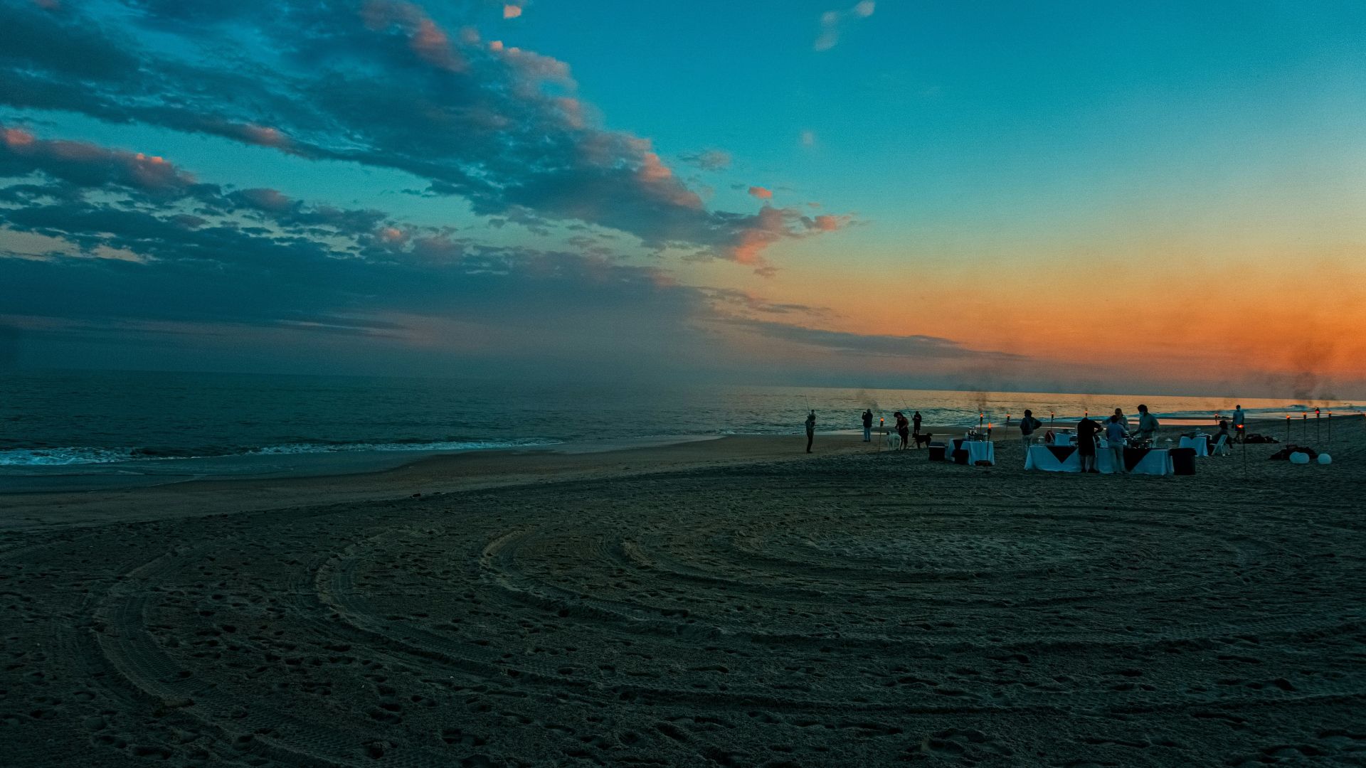 A wide view of a sandy ocean beach at sunset, featuring a rich orange and blue sky, dark waves, and a small gathering of people with tables set up for a special event on the sand.