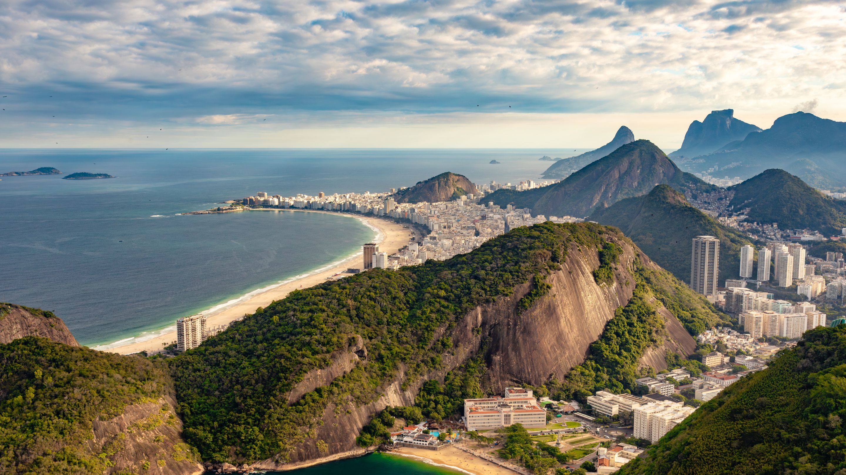 An elevated, wide view of the expansive, crescent-shaped Copacabana Beach and the dense city of Rio de Janeiro, with the Atlantic Ocean on the left and the iconic Sugarloaf Mountain and other steep, green mountains visible in the distance under a partly cloudy sky.