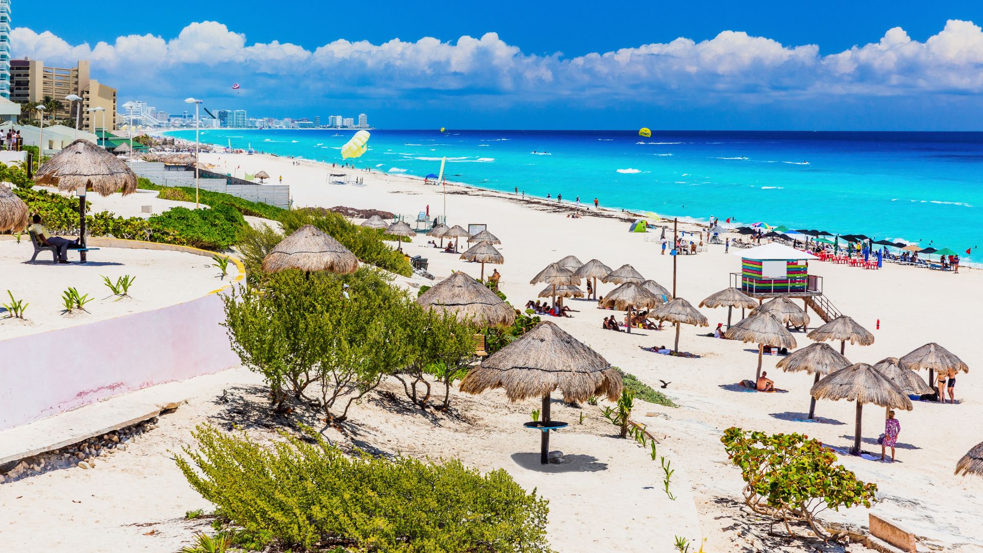 A bright, sunny view of the wide, white sandy expanse of Delfines Beach in Cancun, Mexico. Several traditional palapa umbrellas dot the sand, overlooking the vibrant turquoise and blue ocean waters. A multi-story hotel and urban skyline are visible in the distance.