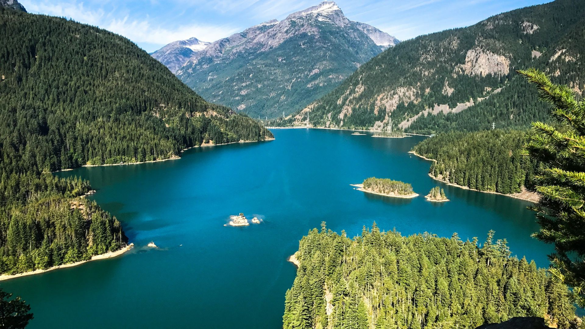A panoramic daytime photograph of the vibrant, turquoise-colored Diablo Lake, nestled among steep mountainsides covered in dense, dark green coniferous forests. Several small, tree-covered islets dot the center of the lake. A rugged, snow-capped peak rises in the distance under a bright blue sky with wispy white clouds.