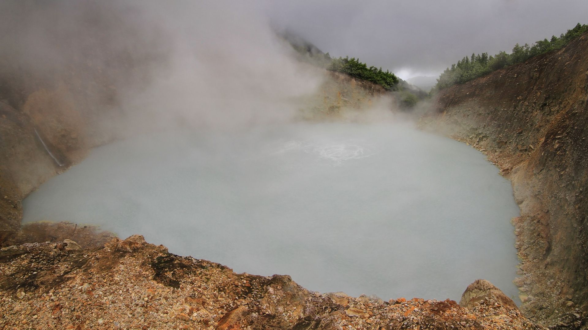 An atmospheric photograph of the second-largest boiling lake in the world, showing a large pool of bubbling greyish-blue water constantly enveloped in thick white steam and surrounded by a barren, rugged, rocky volcanic landscape under a cloudy sky.