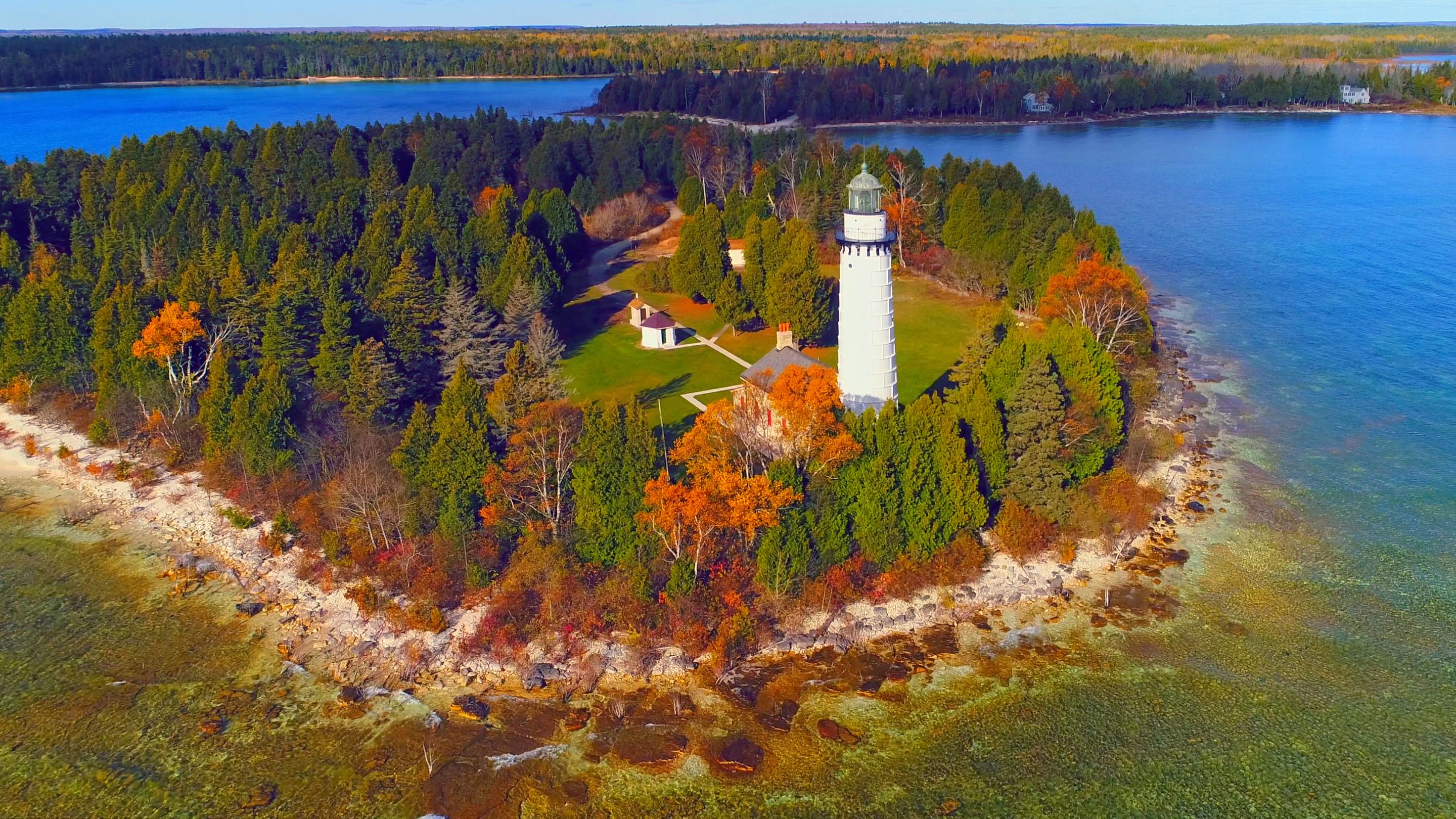 An aerial view of the Cana Island Lighthouse, an 89-foot tall white tower connected to a keeper's house on a small, tree-covered island, with a rocky shoreline and the blue water of Lake Michigan surrounding it.