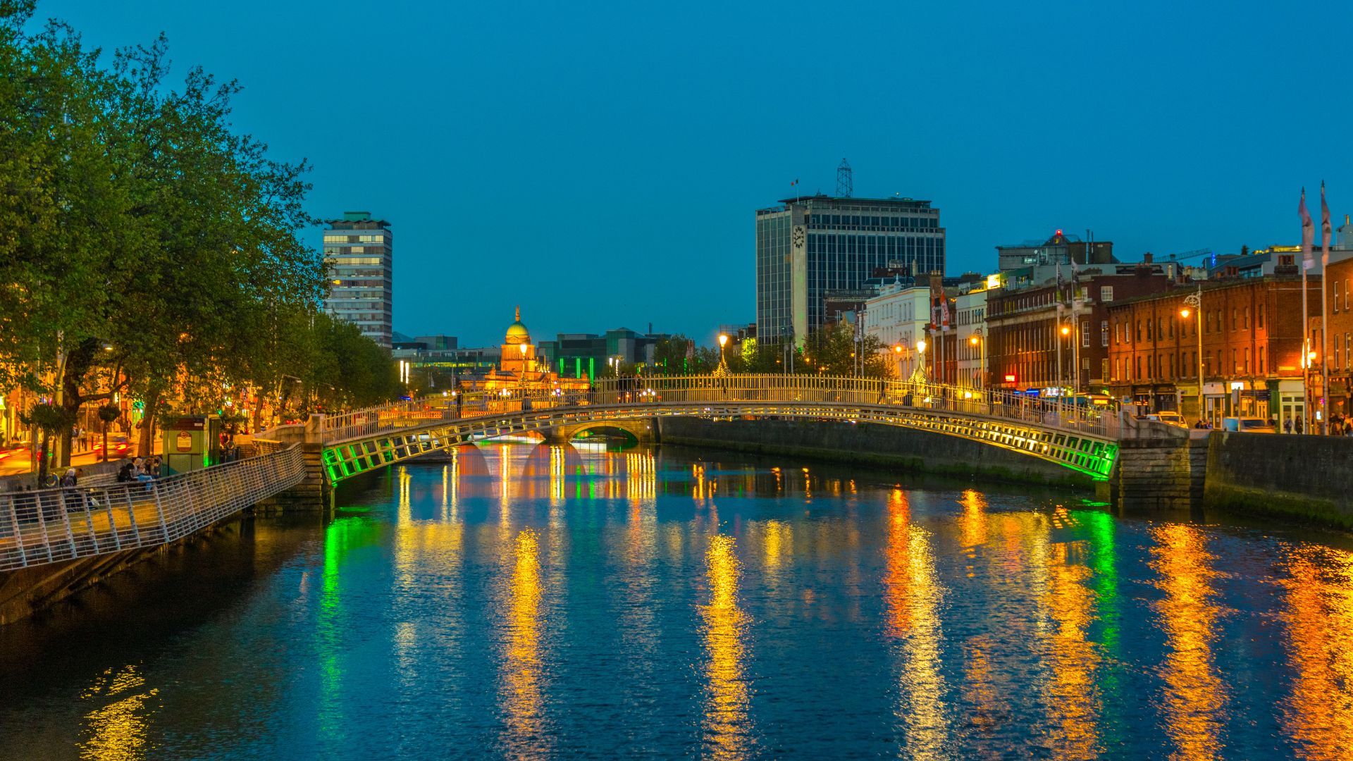 A nighttime view of the iconic white, cast-iron Ha'penny Bridge in Dublin, Ireland, with its architectural arches illuminated in green light, reflecting on the surface of the River Liffey amidst the warm orange glow of surrounding city buildings.