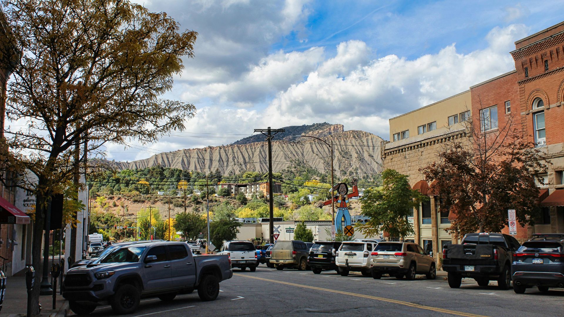 A landscape photograph of a downtown street lined with cars and buildings, framed by autumn trees. In the background, a large, rugged mountain with distinctive horizontal rock layers rises dramatically under a cloudy blue sky. A power pole in the mid-ground features a small Native American-themed mural or decoration.
