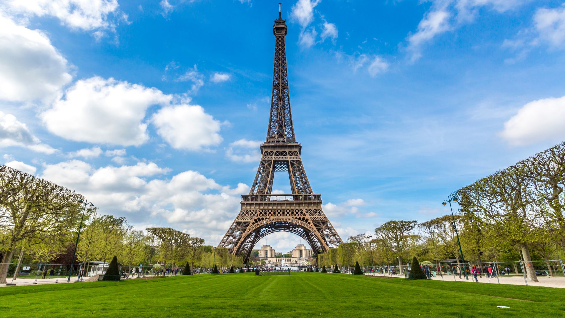 The iconic Eiffel Tower stands tall against a bright blue sky with white clouds, as seen from the green lawns of the Champ de Mars in Paris.