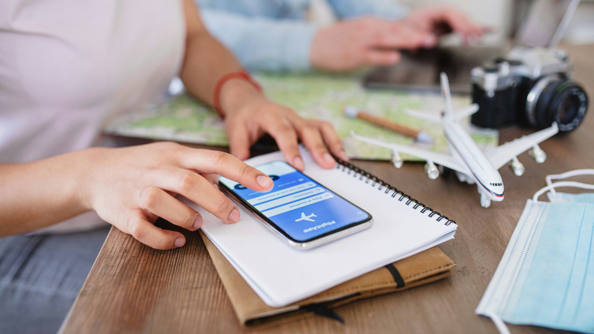 A stock photograph of two people at a wooden table using a smartphone and laptop to plan a trip, with a toy airplane, a map, a camera, and face masks nearby.