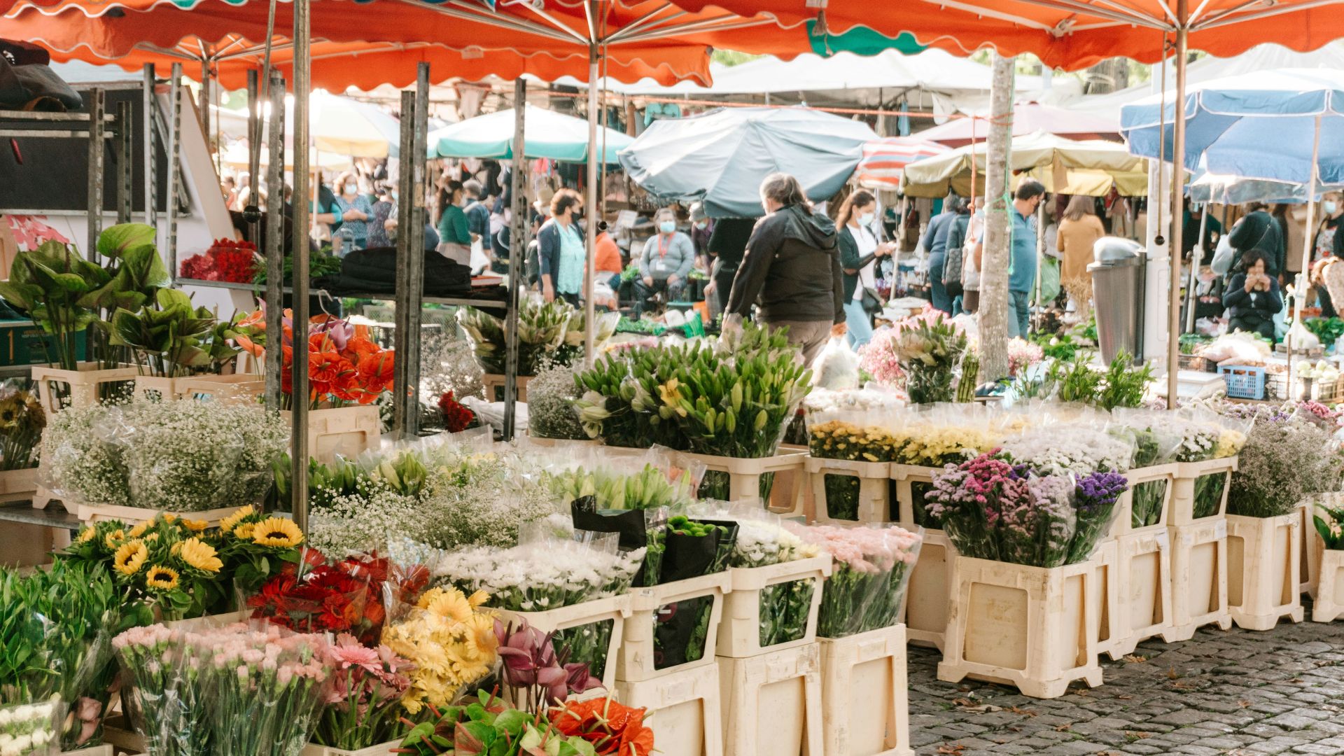 A bustling daytime outdoor flower market on a cobblestone street, possibly in Europe. Several stalls with orange awnings display an abundance of fresh, vibrant flowers in white plastic crates, including sunflowers, roses, gypsophila, and various mixed bouquets. People browse the stalls in the background.