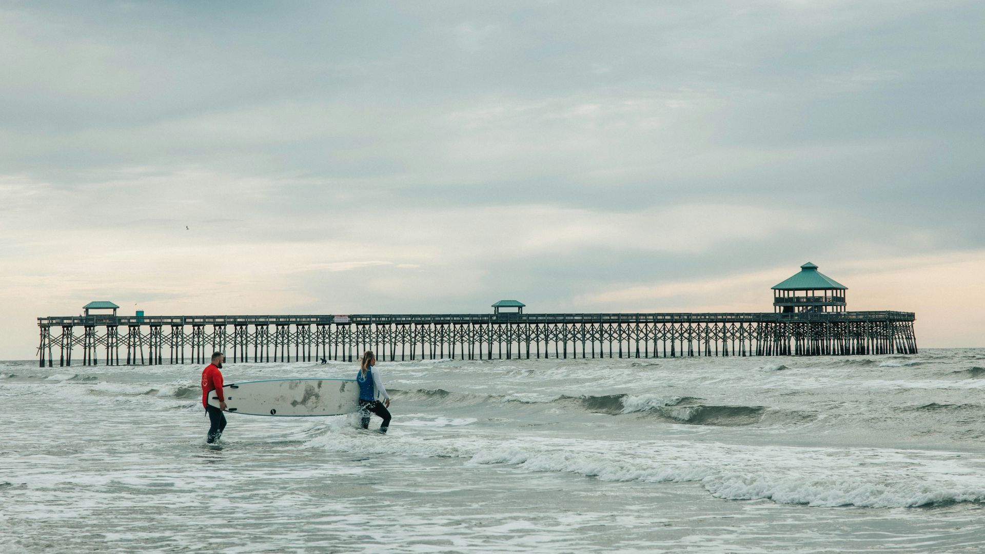 Two people with a surfboard stand in the shallow, wavy ocean water in the foreground, with a long, wooden fishing pier featuring several green-roofed gazebos extending into the Atlantic Ocean under an overcast sky.