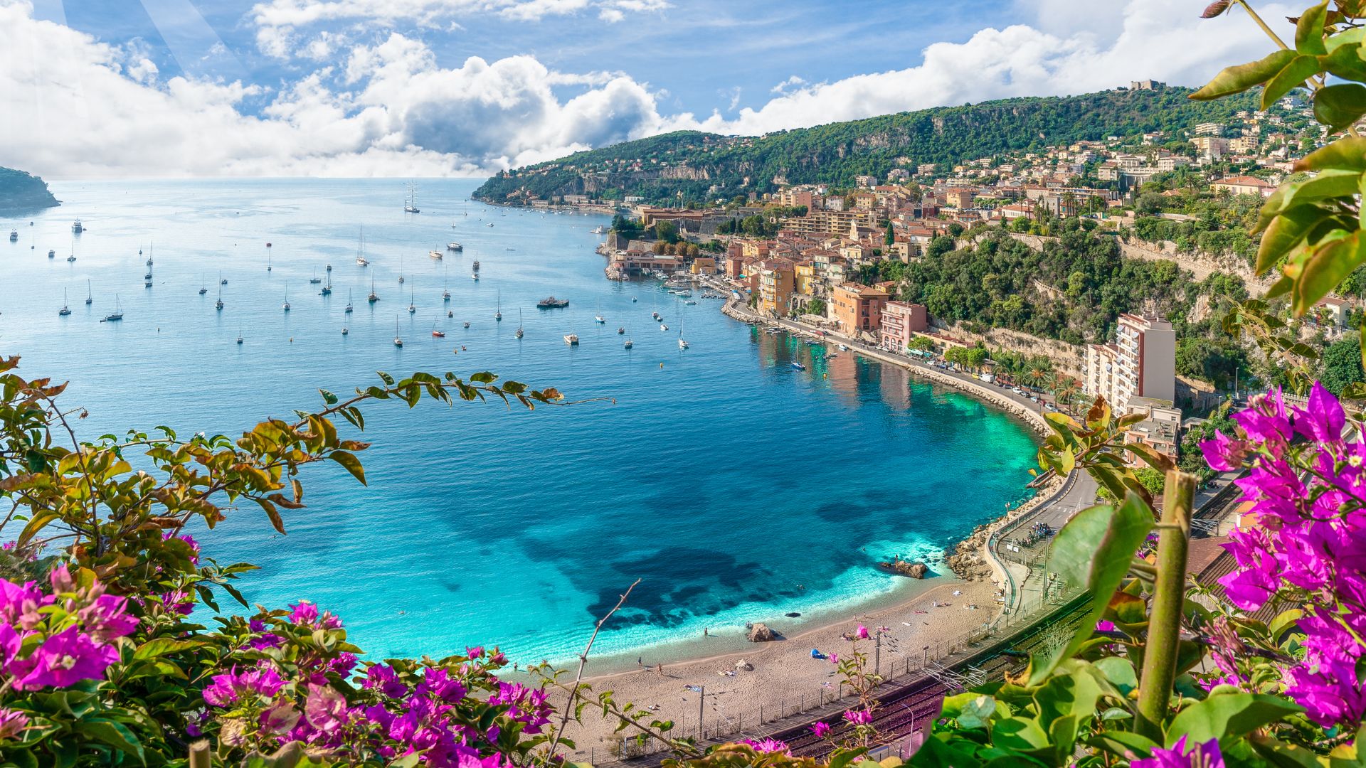 A scenic, elevated view of the turquoise Mediterranean waters and the curved beach of Villefranche-sur-Mer in the French Riviera, lined with colourful buildings and framed by lush green hillsides and bright purple flowers.
