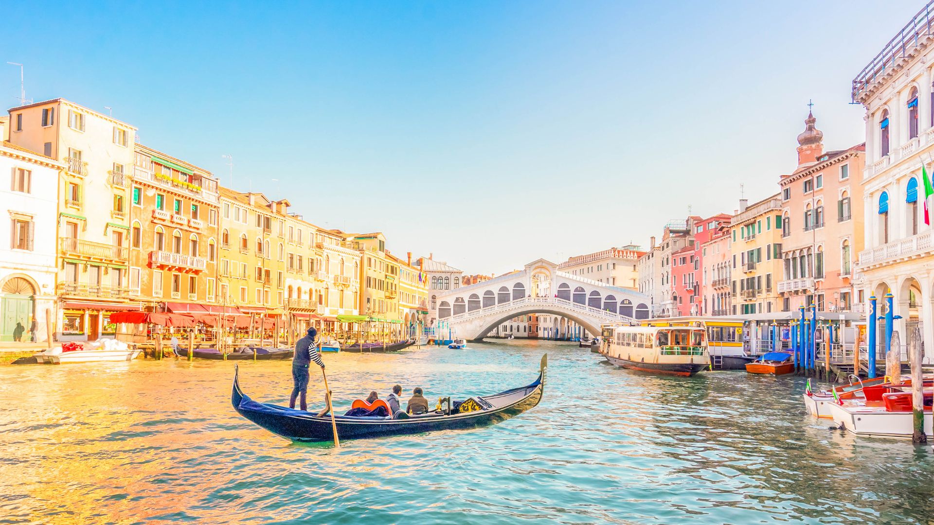 A classic, sunlit view of the Rialto Bridge and the Grand Canal in Venice, Italy, showing a dark blue gondola in the foreground with passengers, surrounded by historic buildings.