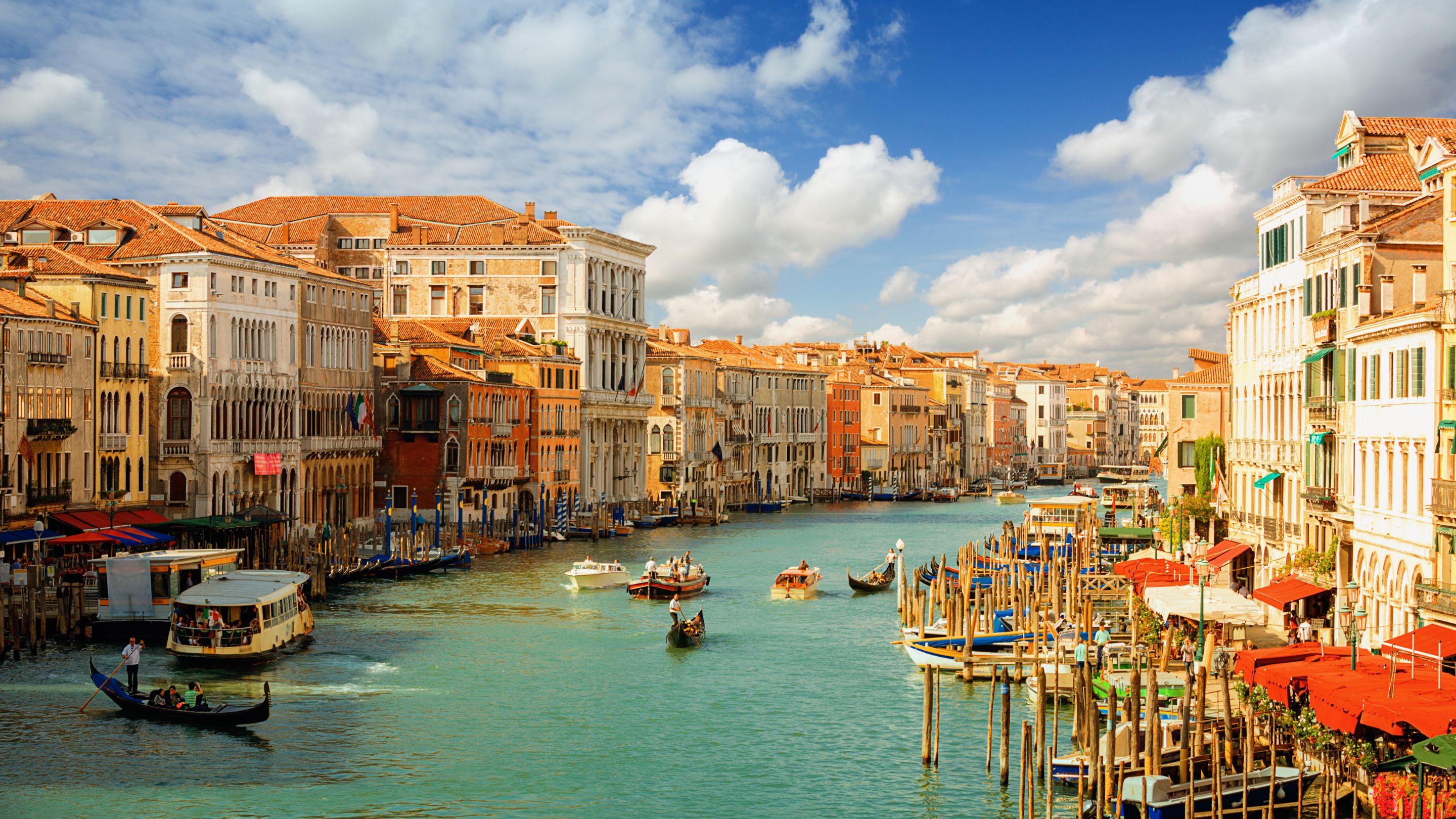 A daytime view of the busy Grand Canal in Venice, Italy, lined with colorful historic buildings and numerous traditional gondolas and water taxis navigating the turquoise water under a blue sky with white clouds.