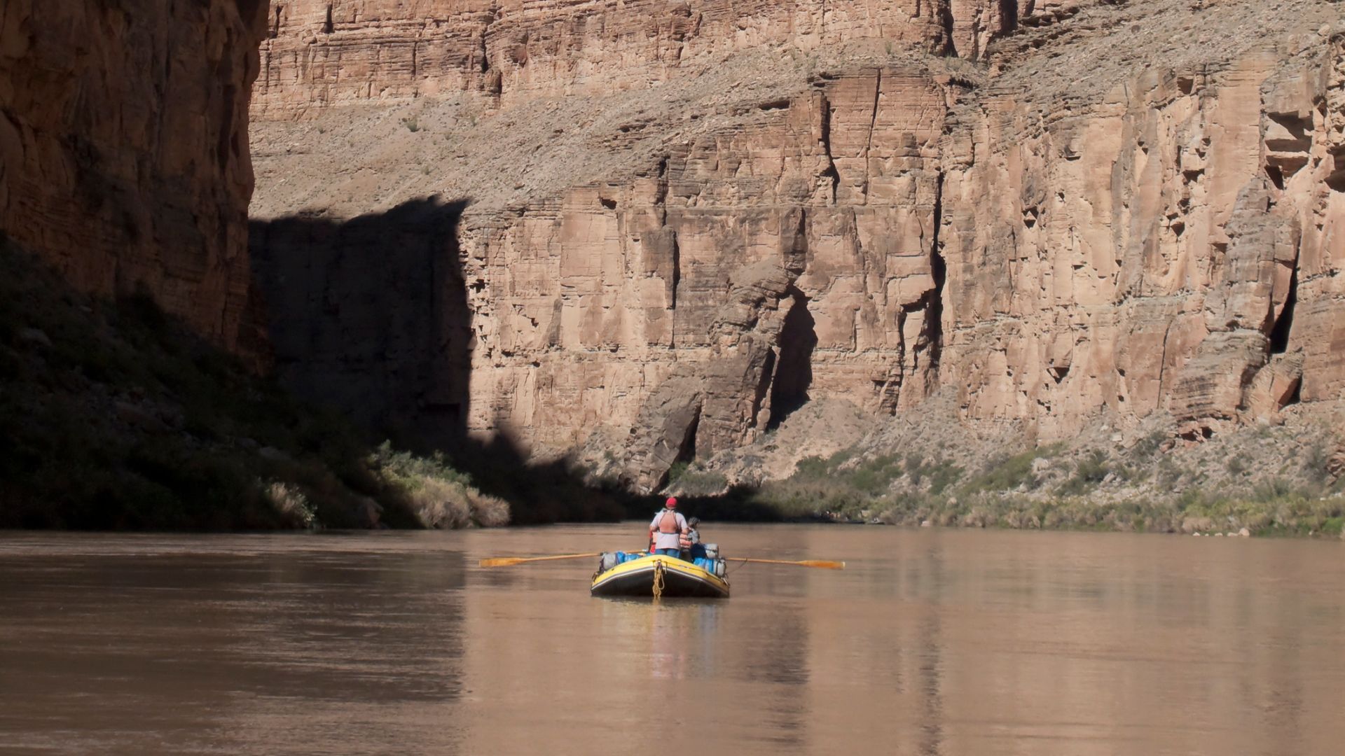 A single person in a yellow inflatable raft floats on the wide, muddy Colorado River at the bottom of the Grand Canyon, surrounded by towering, steep rock canyon walls under a sunny sky.