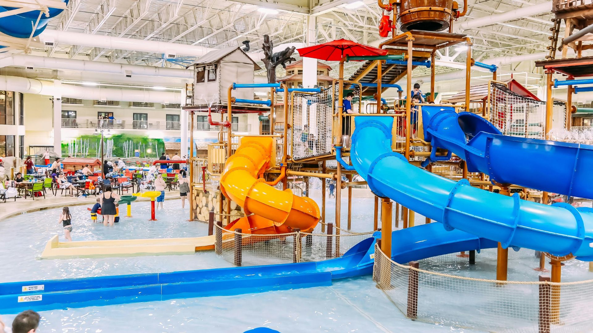 An indoor water park facility featuring a large multi-level play structure with orange and blue water slides, a giant tipping bucket, and a shallow pool area with people in it.