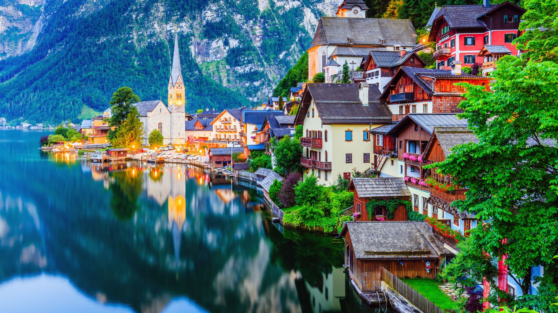 A scenic, twilight view of the picturesque village of Hallstatt, Austria, nestled between steep green mountains and a still lake that reflects the illuminated houses and church spire.