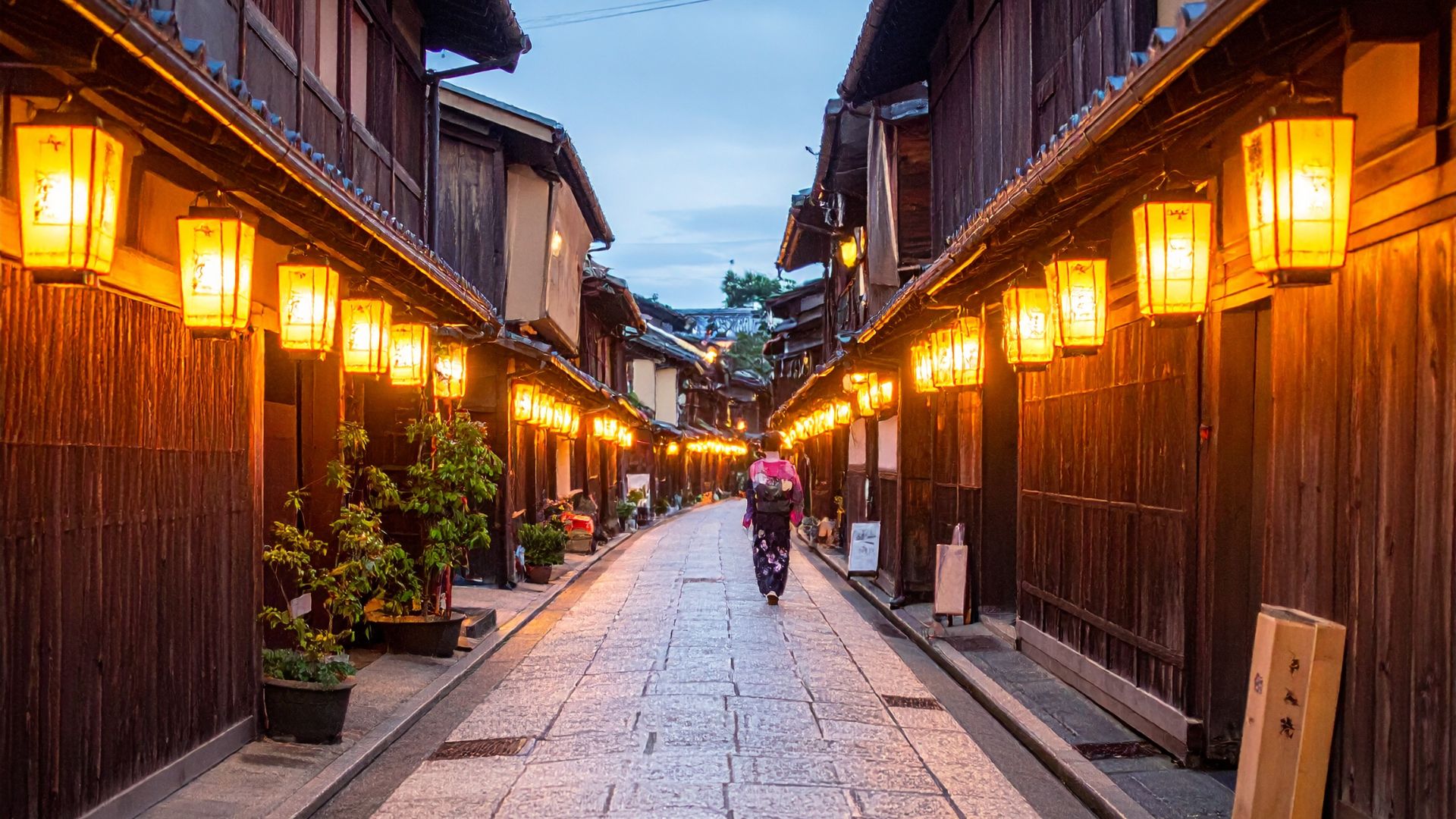 An atmospheric photo of a narrow, traditional Japanese alleyway in Kyoto's Gion district, lined with wooden buildings and illuminated paper lanterns at twilight.
