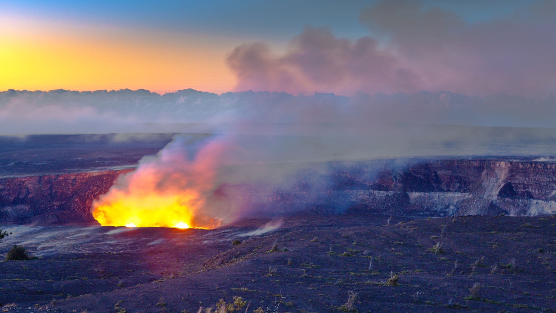A stunning panoramic photograph of the actively erupting Kīlauea volcano in Hawaiʻi Volcanoes National Park. The image captures a bright, glowing lake of molten lava deep within the Halemaʻumaʻu crater at twilight, contrasting sharply with the dark, rugged volcanic landscape and the pastel hues of the sky. Steam and volcanic gases billow from the crater, adding drama to the scene.