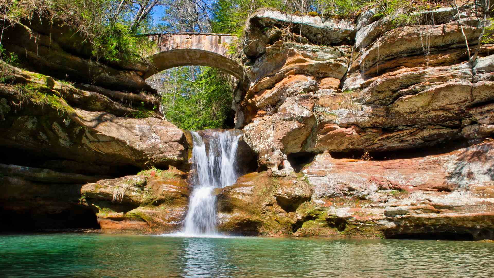 A photograph of the Upper Falls waterfall cascading into a clear, turquoise pool at Hocking Hills State Park in Ohio. The falls are surrounded by mossy, layered sandstone cliffs and rock formations, with a rustic stone arch bridge visible spanning the gorge overhead.