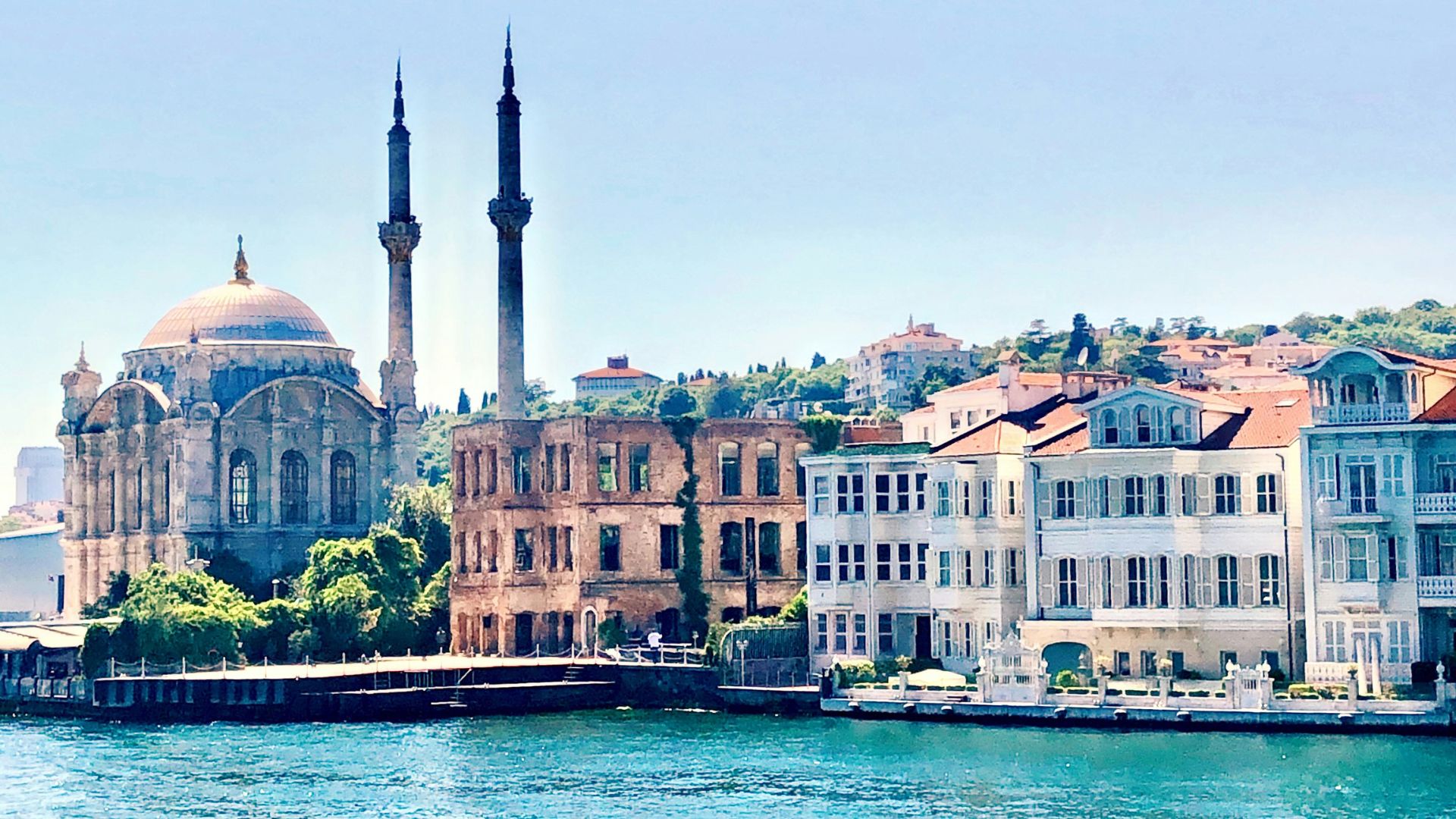 A scenic, daytime photograph of the ornate, Neo-Baroque style Ortaköy Mosque and classic Ottoman-era wooden houses ("yalı") situated on the edge of the vibrant blue Bosphorus Strait in Istanbul, under a clear sky.