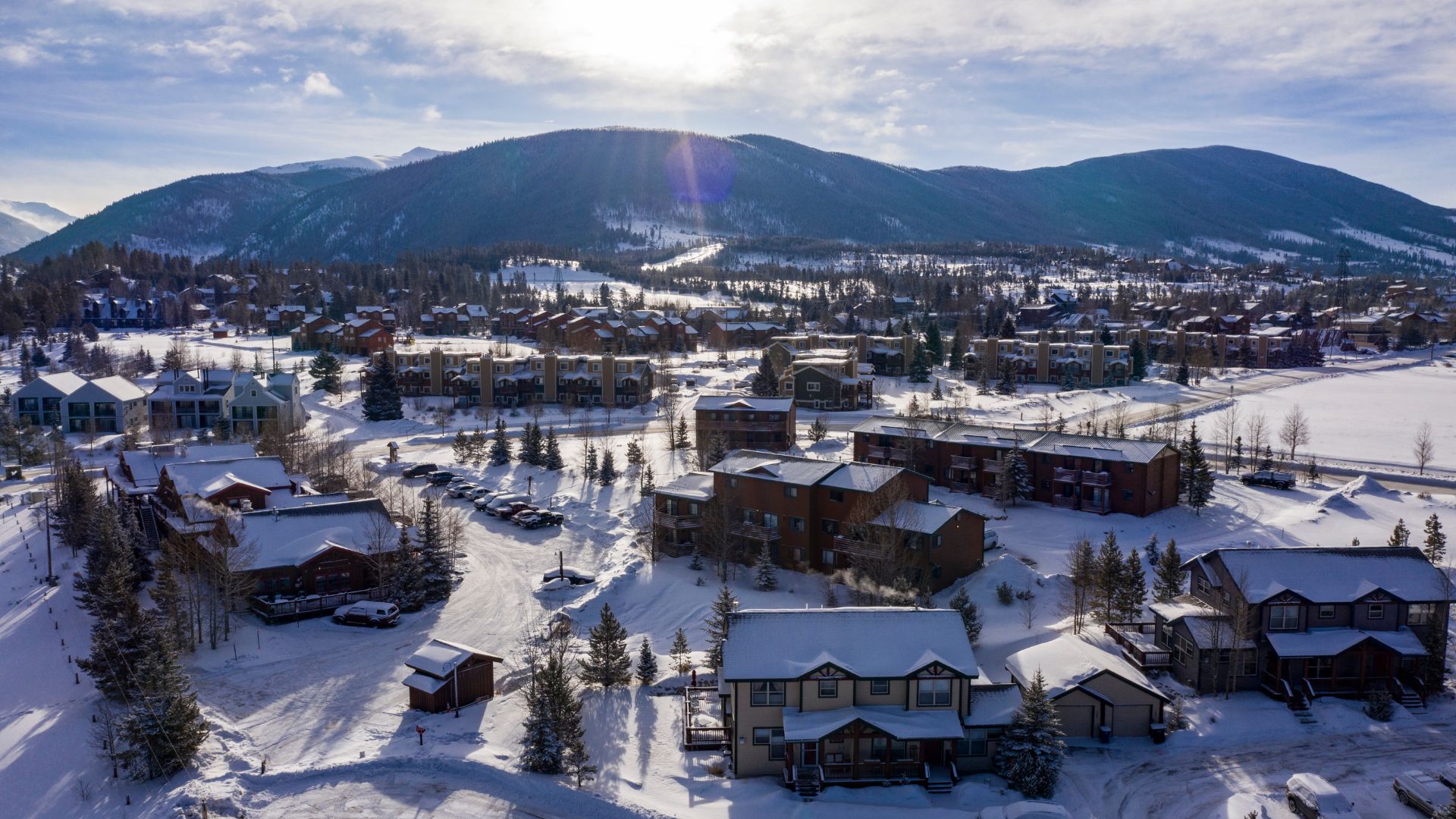An aerial view of a snow-covered mountain resort town with many houses and lodges at the base of a large, snowy mountain.